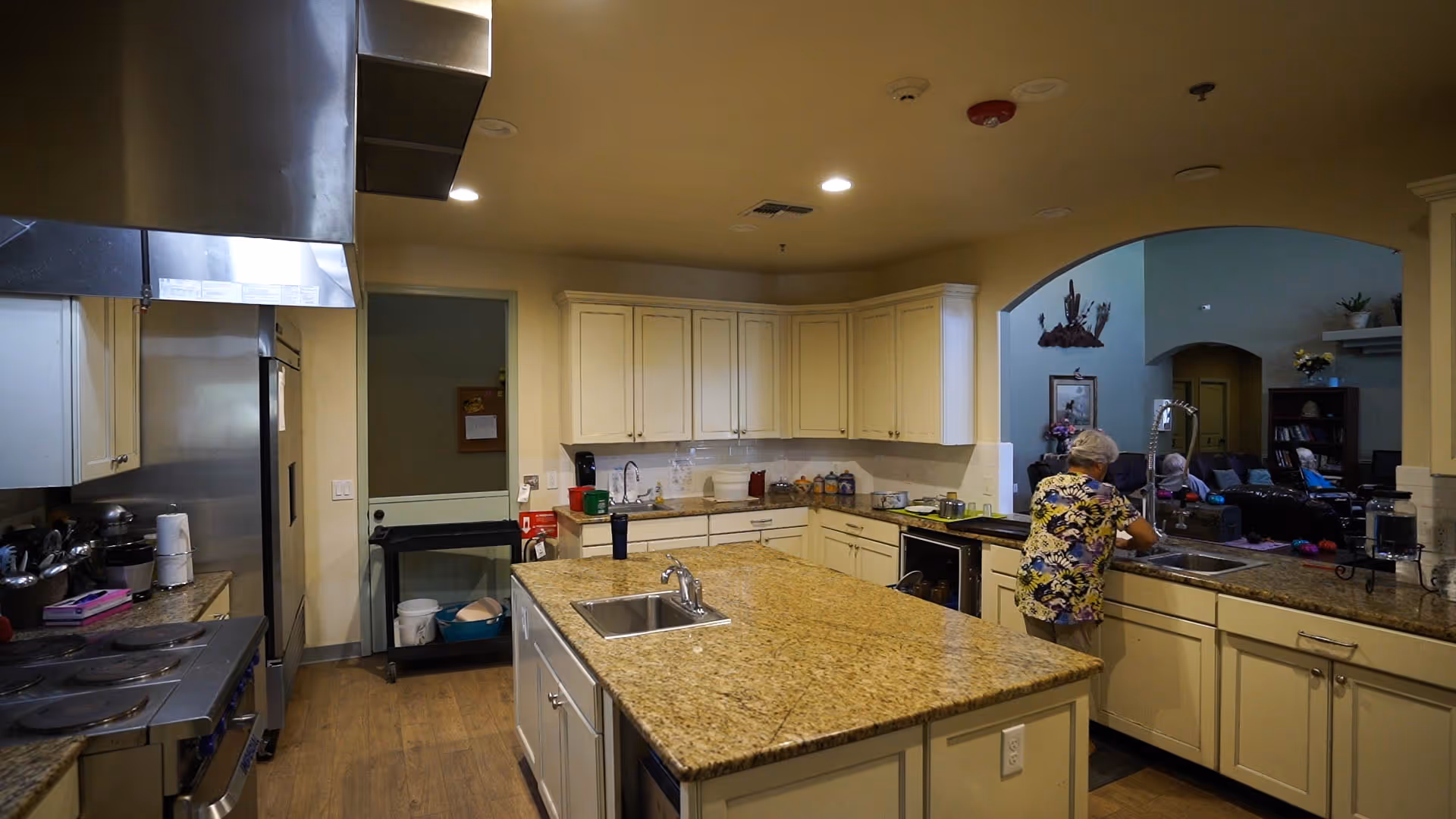 A spacious kitchen with cream-colored cabinets and granite countertops. There is a large island in the center with a sink. A woman is standing at the sink on the right side, washing or preparing something. The kitchen opens into a living room area where several elderly people are seated on couches. The floor is wooden, and the kitchen has stainless steel appliances including a stove and refrigerator.