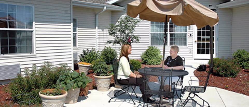 Two women sitting and talking at a round metal table with an umbrella on a patio outside a building with white siding. There are several potted plants and shrubs around the patio area.
