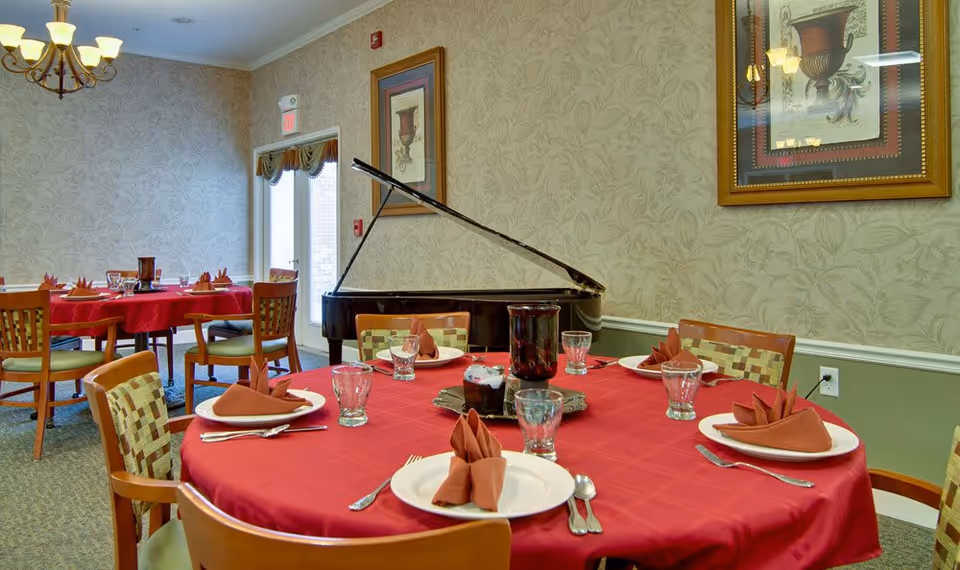 Dining room with round tables covered in red tablecloths, each set with plates, folded napkins, glasses, and silverware. A grand piano is positioned against a wallpapered wall with framed artwork. The room is softly lit by a chandelier and natural light from a door with a window.