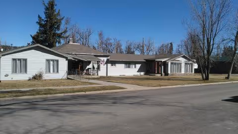 Single-story white building with multiple sections and a gray roof, situated along a paved street with leafless trees and a clear blue sky in the background.
