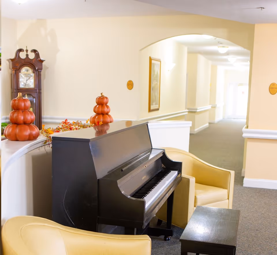 A cozy indoor seating area with a black upright piano, two yellow armchairs, and a small black table. The background shows a hallway with cream-colored walls, a grandfather clock, framed artwork, and autumn-themed decorations including stacked pumpkin ornaments.
