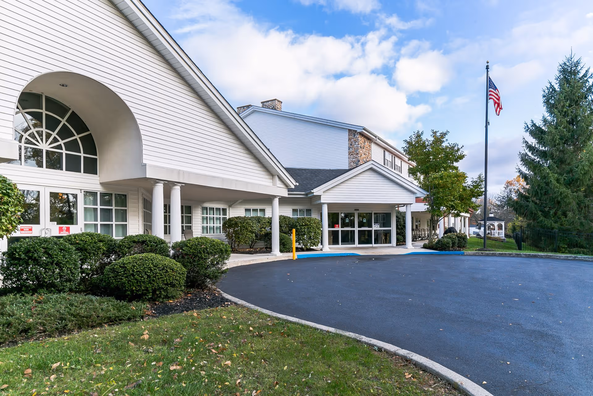 Exterior view of a senior living facility building with white siding, large windows, and a covered entrance supported by white columns. There are neatly trimmed bushes and a green lawn in front, a paved driveway, and an American flag on a flagpole to the right. The sky is partly cloudy.