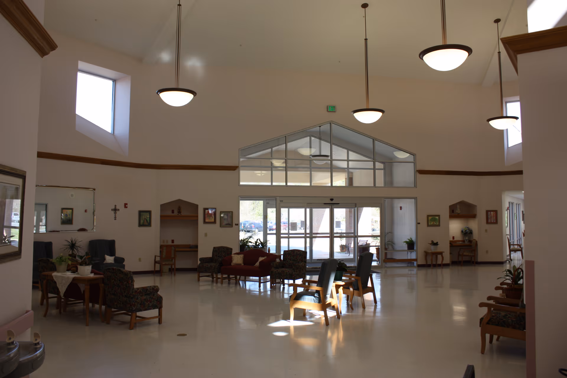 Spacious and well-lit common area in a senior living facility with high ceilings, large windows, and multiple seating arrangements including armchairs and sofas. The room features hanging pendant lights, plants on tables, framed artwork on the walls, and glass doors leading outside.