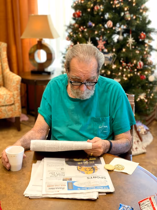 An elderly man wearing glasses and a green t-shirt is sitting at a round table reading a newspaper. He holds a white cup in one hand. On the table are more newspapers, a napkin with two cookies, and some snack wrappers. Behind him is a decorated Christmas tree and a lamp on a side table next to an upholstered chair.