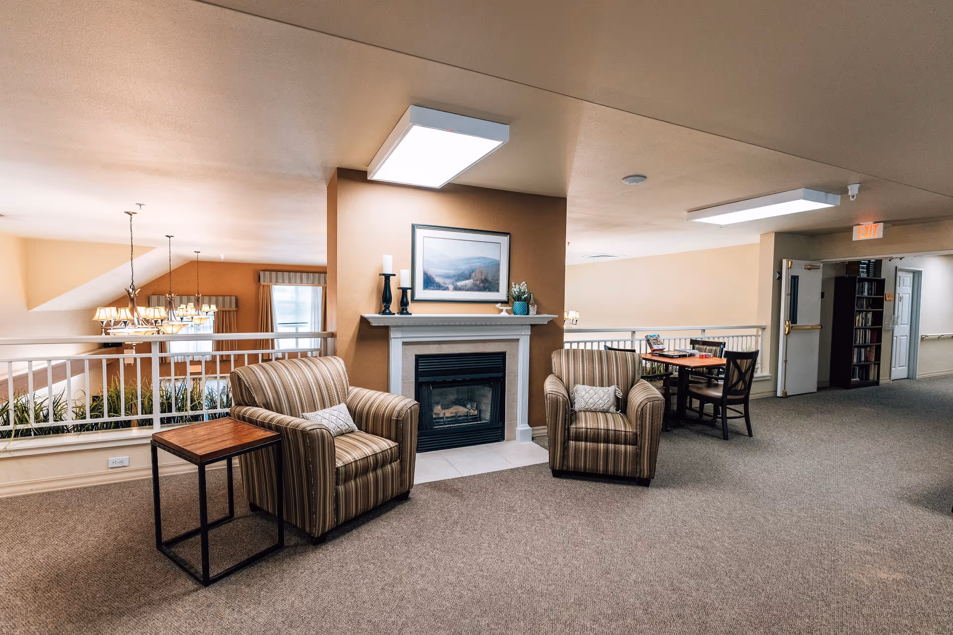 A cozy indoor common area with two striped armchairs and a small wooden side table in front of a fireplace. Above the fireplace is a framed landscape painting and decorative items. In the background, there is a dining table with chairs and a bookshelf near an open door. The space is well-lit with ceiling lights and has beige walls and carpeted floors.