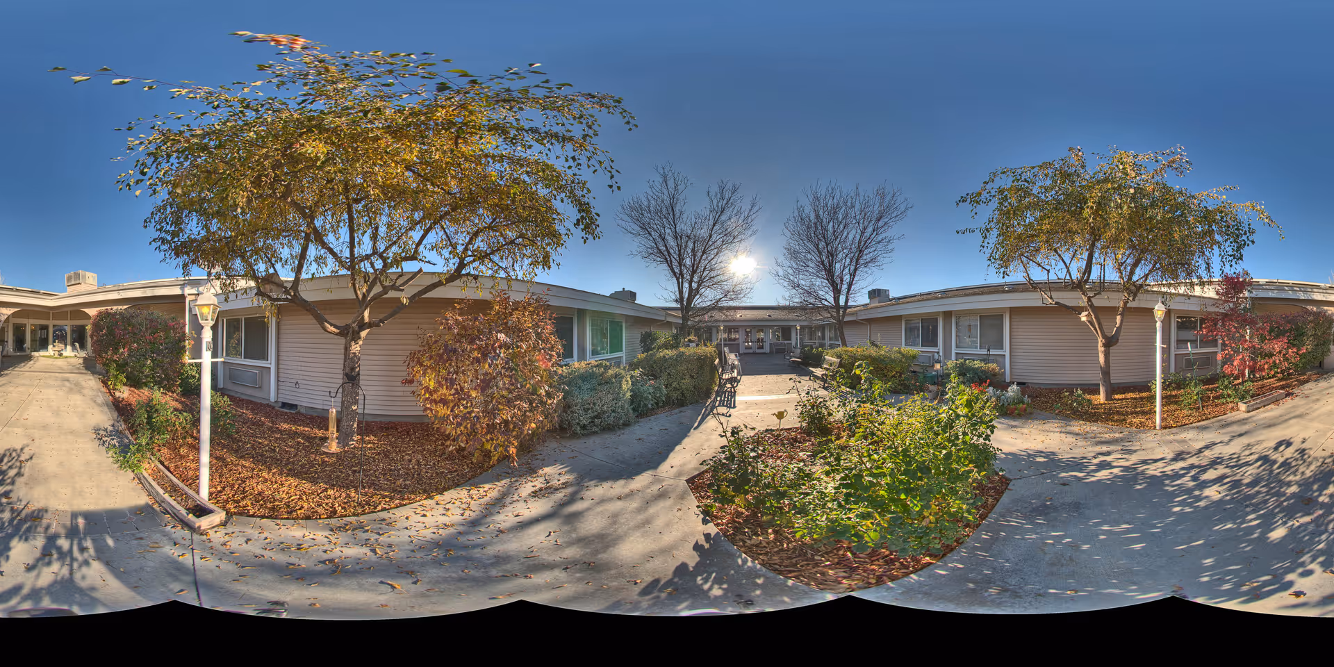 Sunny landscaped courtyard and entrance of a single-story assisted living building with walkways, benches, trees, and shrubs.