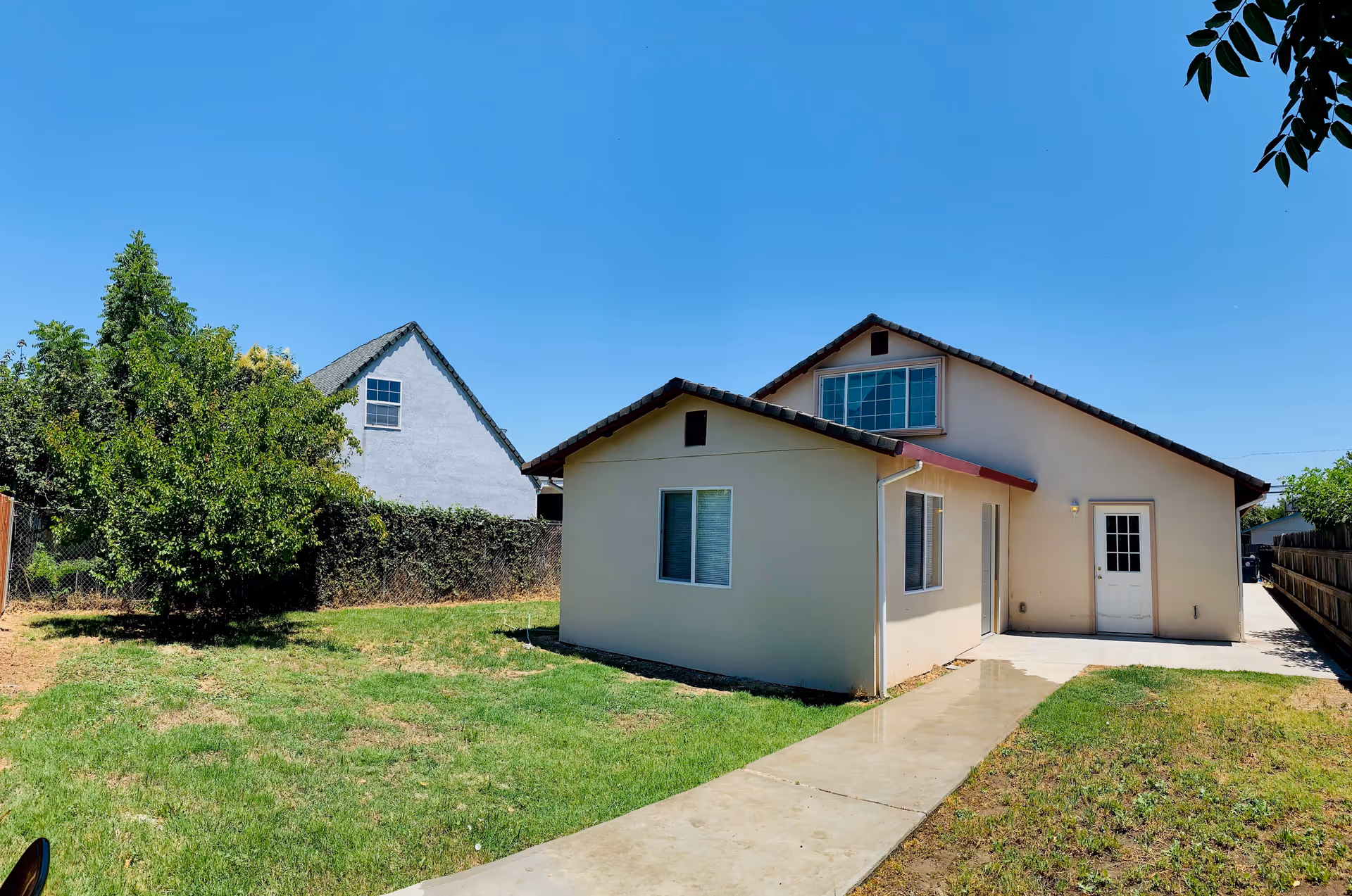 Exterior view of a single-story beige building with a sloped roof, several windows, and a white door. The building is surrounded by a green lawn with a concrete pathway leading to the door. There are trees and a fence in the background under a clear blue sky.