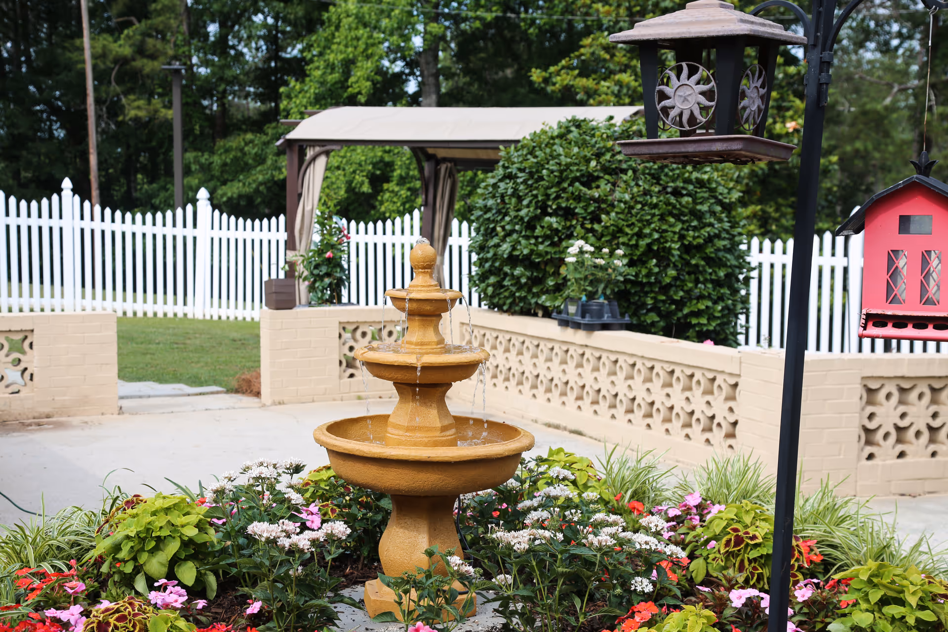 A garden area with a three-tiered water fountain surrounded by colorful flowers and greenery. In the background, there is a white picket fence, a beige brick wall with decorative cutouts, a gazebo with a beige canopy, and a large green bush. A black metal pole holds a decorative lantern and a red birdhouse.