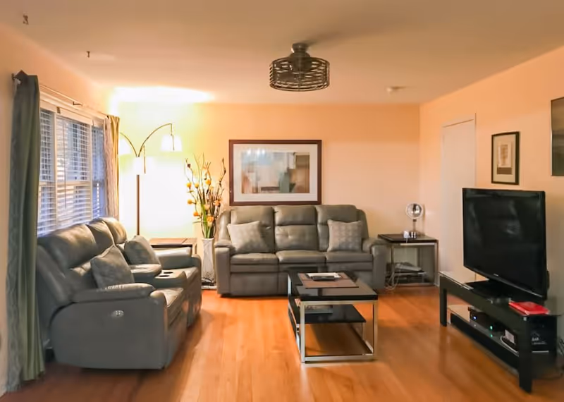 A cozy living room with wooden flooring featuring two gray leather sofas, a coffee table in the center, a flat-screen TV on a stand to the right, a floor lamp with multiple lights near the window, and framed artwork on the wall.