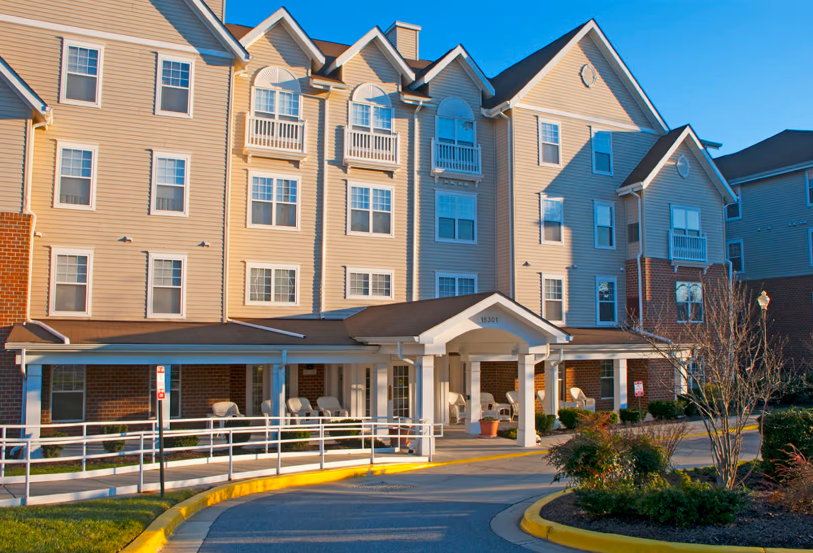 Exterior view of a multi-story senior living facility building with beige siding and brick accents, featuring multiple windows and small balconies. The entrance has a covered porch with white columns and several chairs on the porch. There is a curved driveway with a yellow curb and some landscaping with bushes and small trees in front of the building.