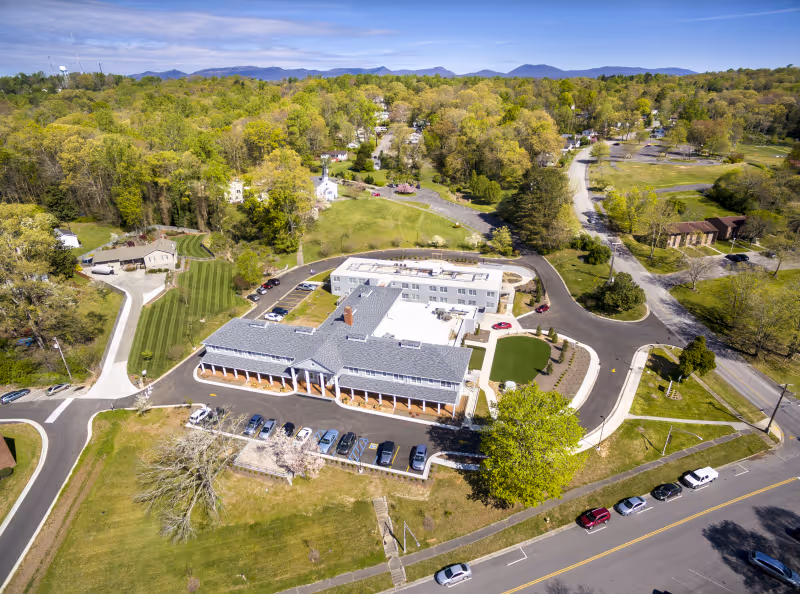 Aerial view of Alexander Guest House facility surrounded by trees and greenery, showing a large building with a parking lot and several cars parked along the road nearby under a clear blue sky.