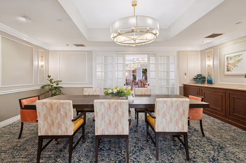 A well-lit dining room with a large rectangular wooden table surrounded by eight upholstered chairs, some with patterned fabric and others in solid orange. A modern chandelier hangs above the table. The room features beige walls with white trim, a patterned carpet, a sideboard with decorative items, and glass-paneled double doors leading to another dining area in the background.