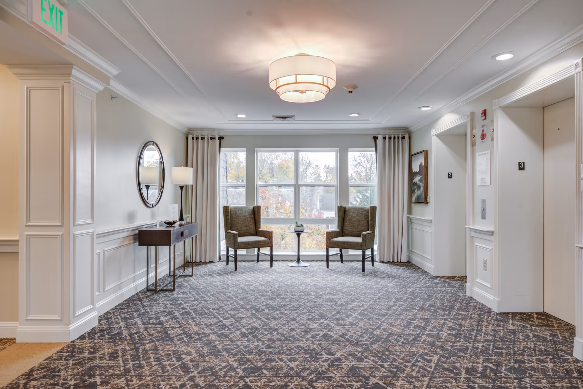 A bright and spacious interior hallway area with two elevators on the right side, two armchairs with a small round table between them in front of large windows with curtains, a console table with a lamp and decorative items on the left wall, and a ceiling light fixture. The carpet has a patterned design and there is an exit sign above a doorway on the left.