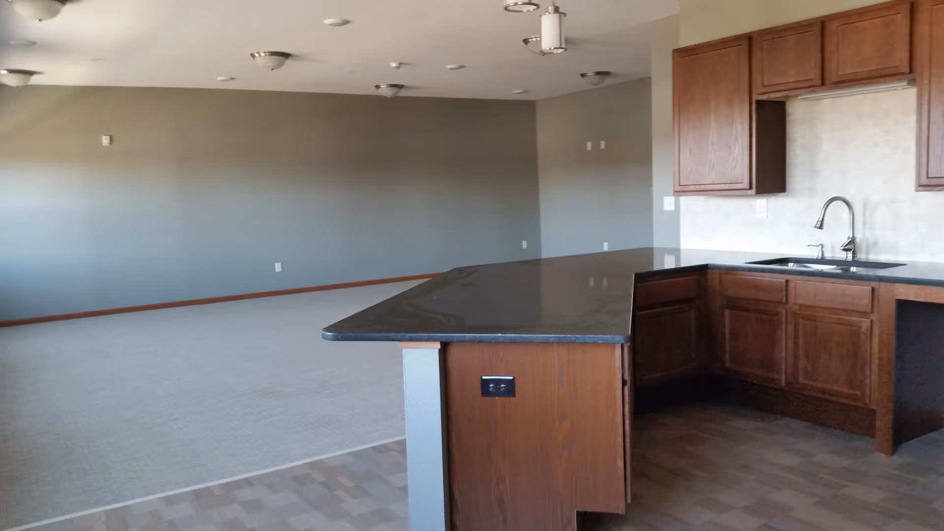 Interior view of a senior living facility showing a kitchen area with wooden cabinets, a black countertop, and a sink with a faucet. Adjacent to the kitchen is an open space with carpeted flooring and gray walls, illuminated by ceiling lights.
