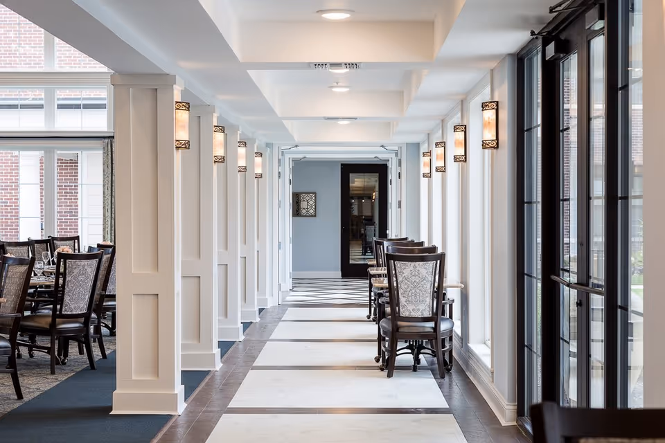 A bright, elegant hallway with white walls and large windows on the right side, featuring decorative wall sconces and a row of wooden chairs with patterned upholstery along the window side. To the left, there is a dining area with tables and chairs visible.