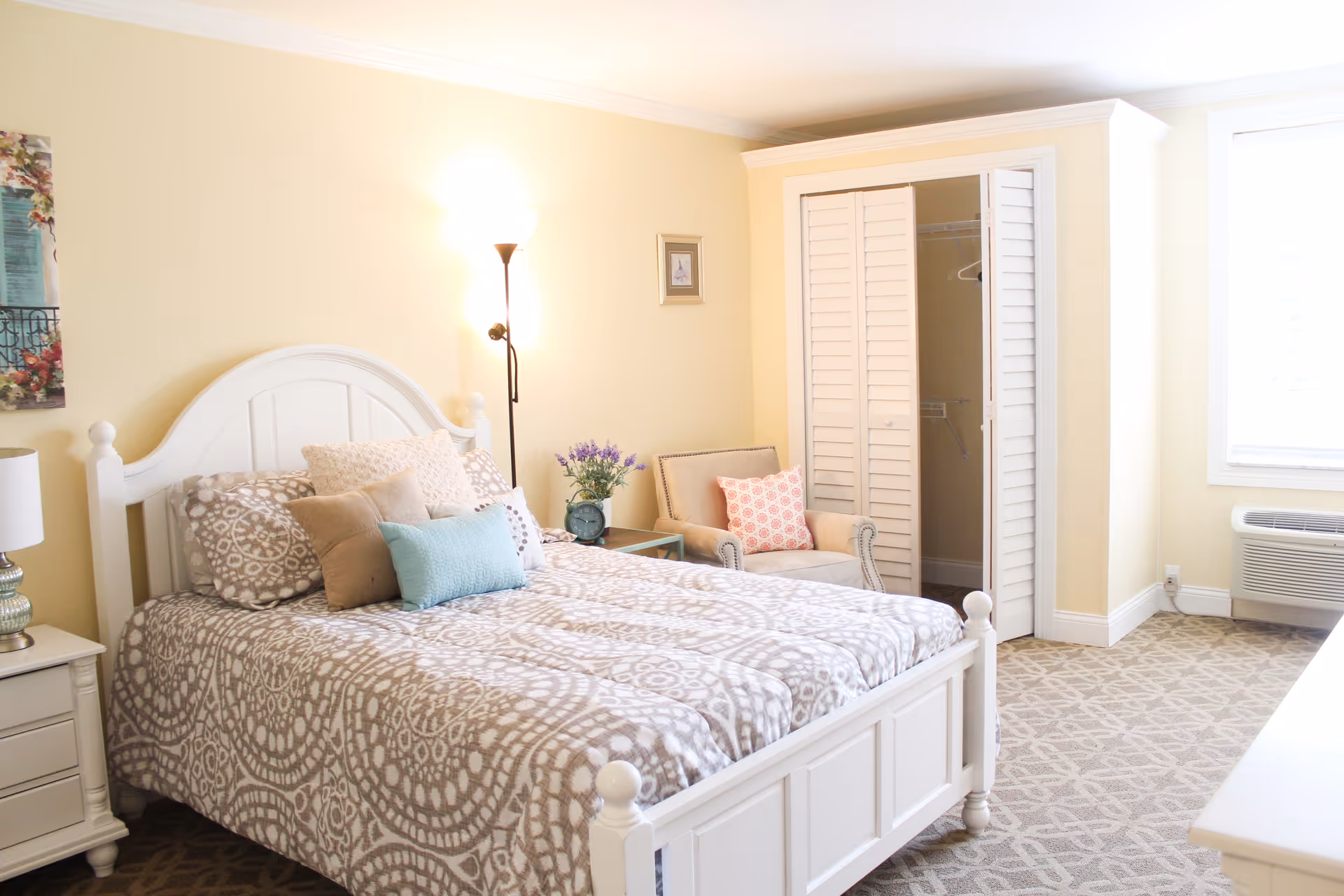 A bright and cozy bedroom with a white wooden bed frame, patterned bedding, and multiple pillows. There is a nightstand with a lamp on the left side of the bed, a floor lamp behind the bed, and a small armchair with a decorative pillow next to a closet with white louvered doors. A window with white blinds and an air conditioning unit is visible on the right side of the room.