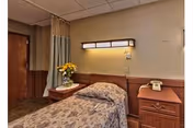 A single bed in a nursing facility room with a floral bedspread, a wooden nightstand with a vase of yellow flowers, a wall-mounted light fixture above the bed, and a wooden cabinet with a vintage telephone on top. The room has beige walls and a curtain partially covering a doorway.