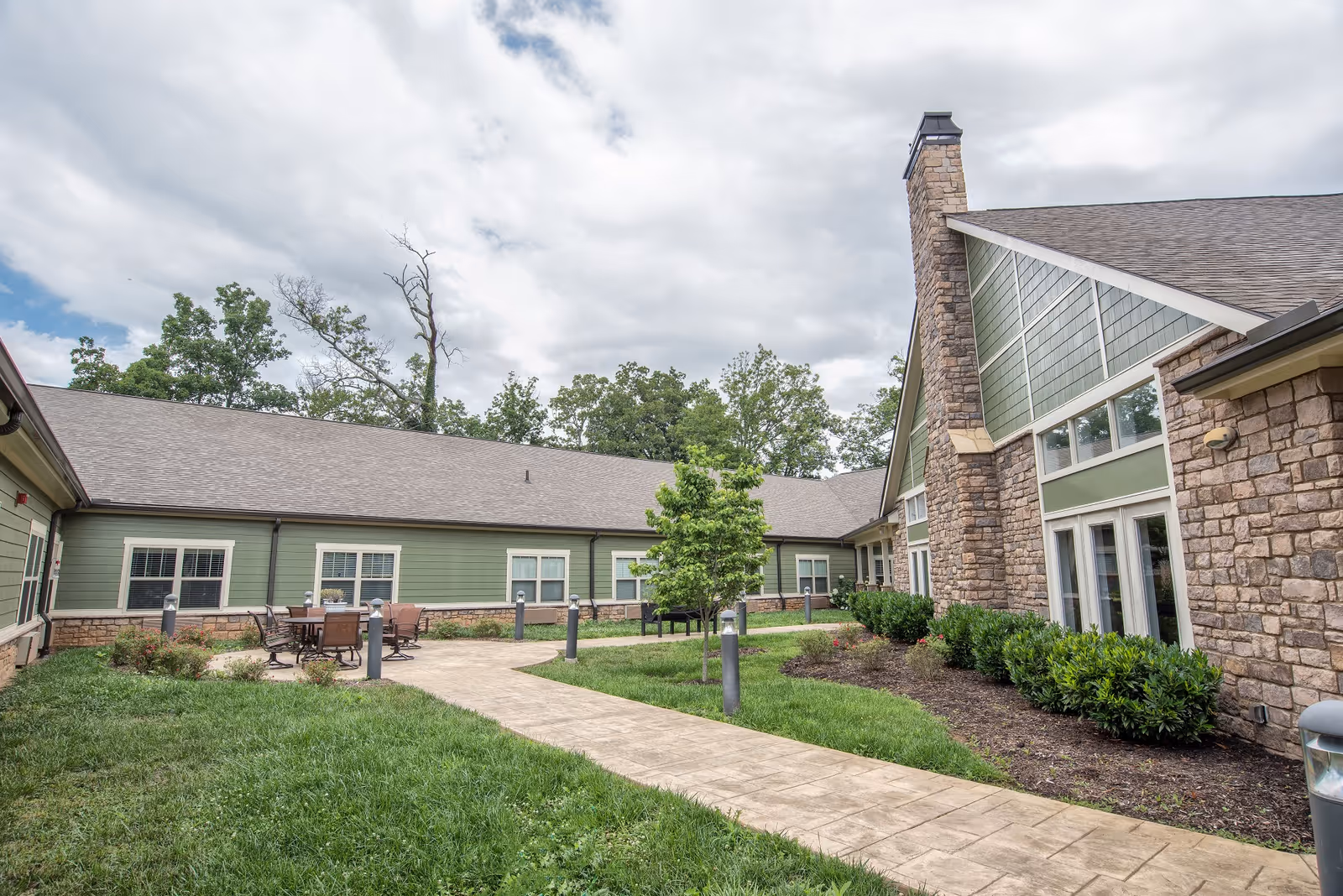 Outdoor courtyard area of Clover Hill Senior Living with a paved walkway, green grass, shrubs, a small tree, and patio furniture. The building exterior features green siding and stone walls with large windows and a chimney under a cloudy sky.
