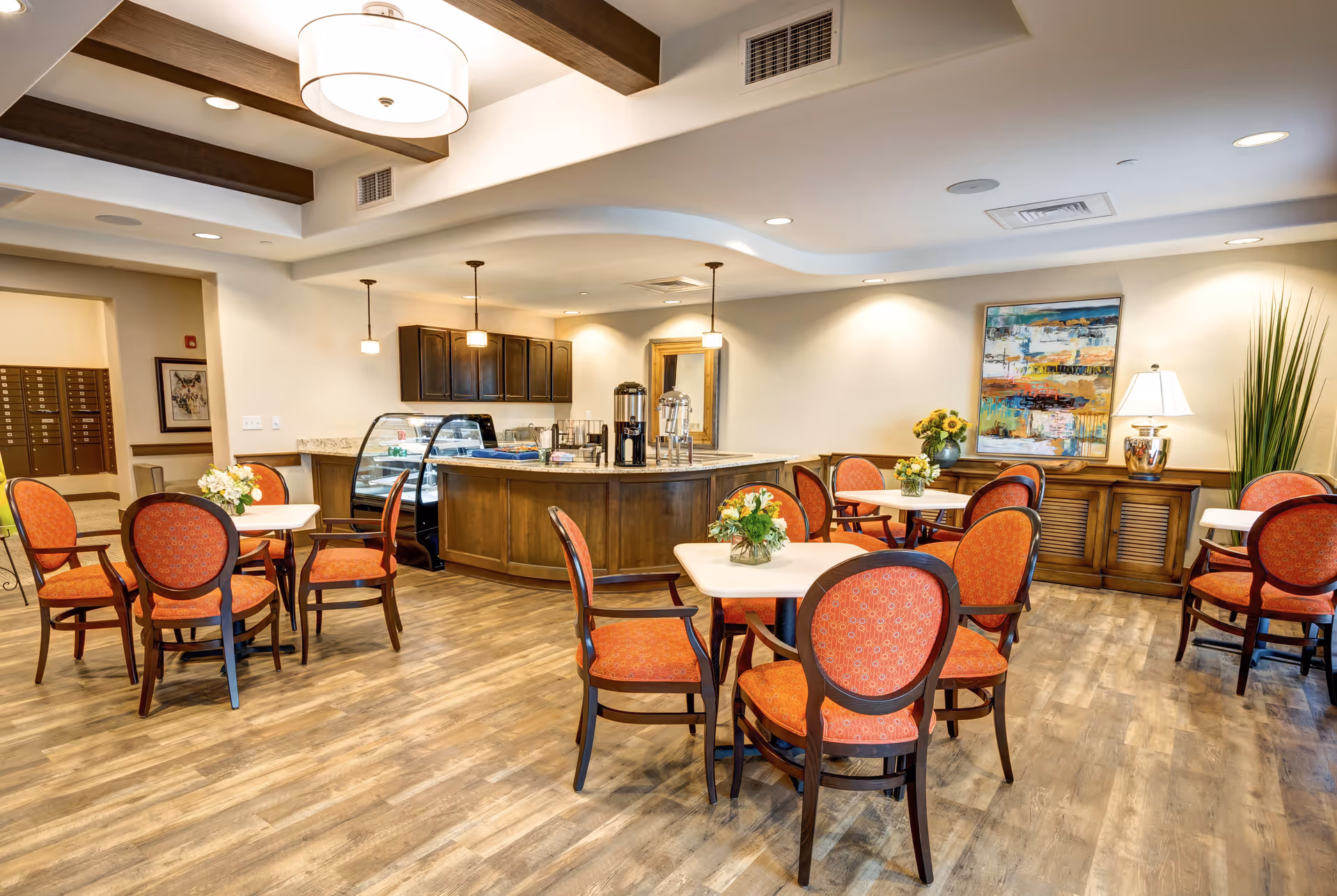 A bright and inviting dining area in a senior living facility featuring several round tables with red cushioned chairs. The room has wooden flooring, a central counter with coffee and beverage dispensers, and a display case. The walls are decorated with a colorful abstract painting, a lamp, and a tall plant. Ceiling lights and wooden beams add warmth to the space.