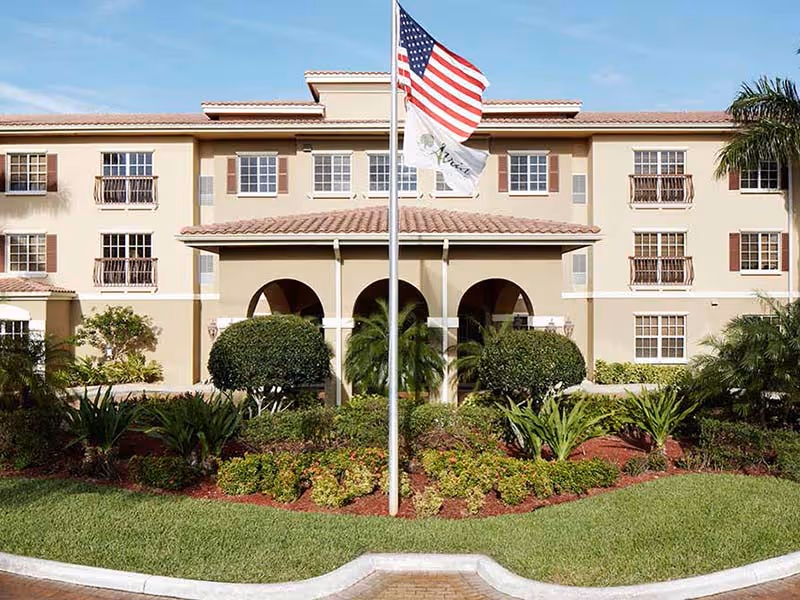 Front exterior view of a three-story senior living facility building with beige walls and a tiled roof. There are multiple windows with brown shutters, a central entrance with three arches, and a flagpole with the American flag and another flag in front. The landscaping includes trimmed bushes, green grass, and various plants.