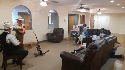 A common area in a senior living facility with several elderly residents seated on leather sofas and chairs. Two men wearing hats are playing guitars near an archway. The room has beige walls, ceiling fans, and recessed lighting.