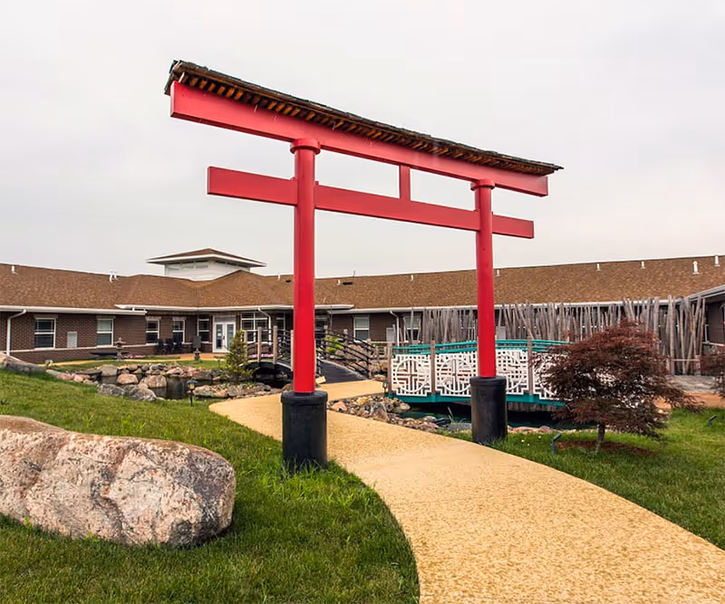 Outdoor view of Addington Place of Des Moines featuring a red torii gate over a curved yellow pathway, green grass, rocks, a small bridge, and a building with a brown roof in the background under a cloudy sky.