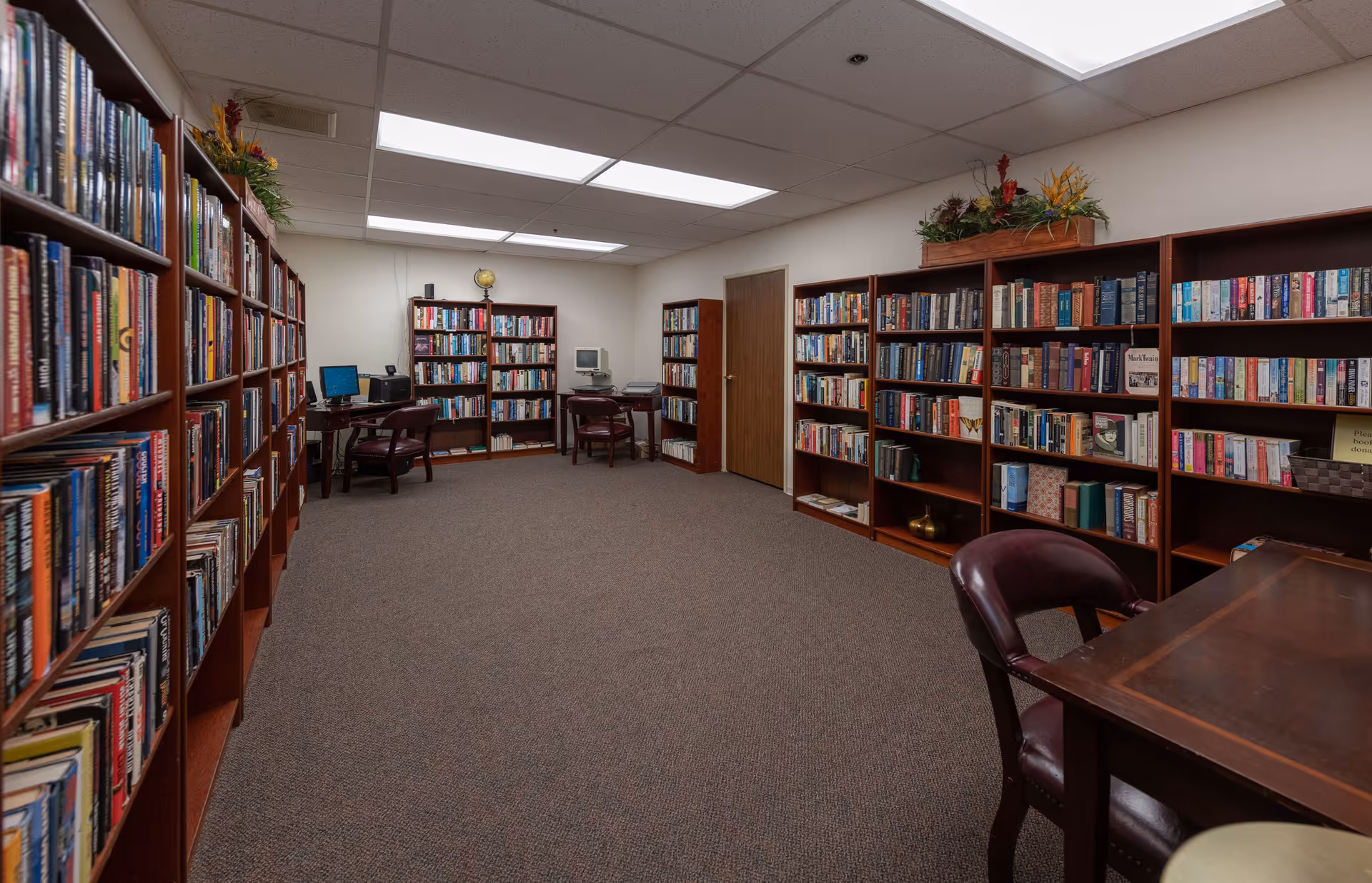 Interior library room with bookshelves lining both walls, tables, chairs and computers.