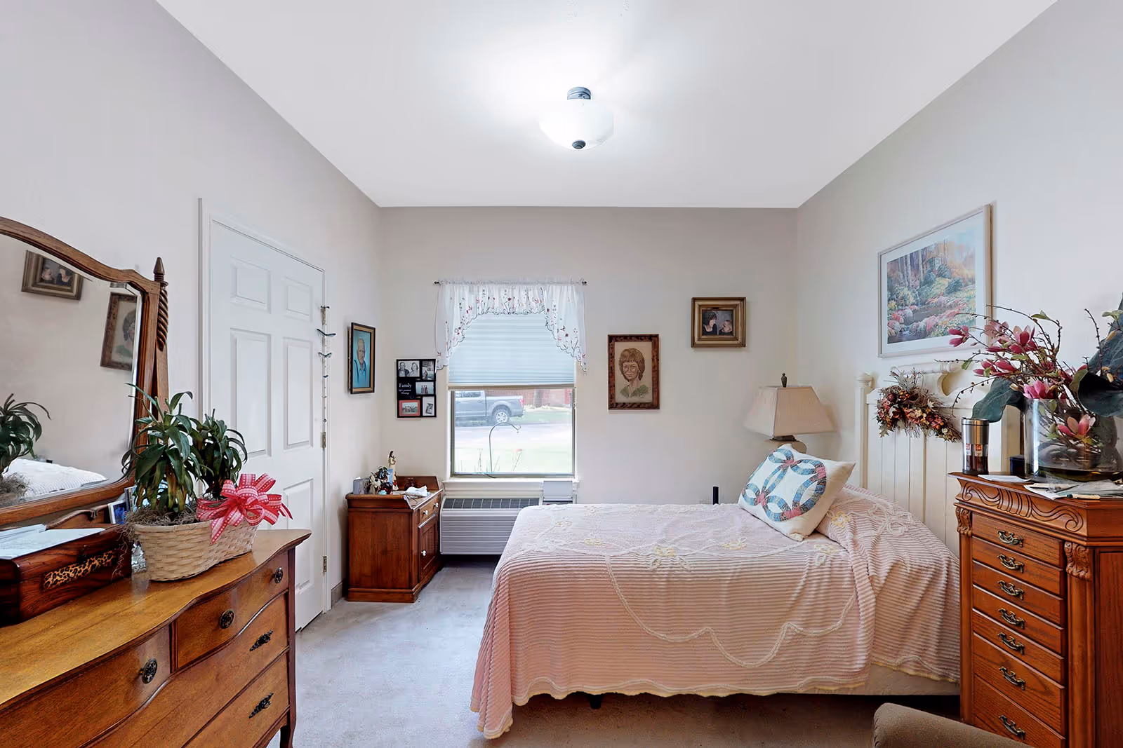 A cozy bedroom with a single bed covered in a light pink bedspread and a decorative pillow. The room has wooden furniture including a dresser with a mirror, a nightstand, and a chest of drawers. There are framed pictures and paintings on the walls, a window with a white curtain, and a ceiling light fixture. A potted plant and some decorative items are placed on the furniture.