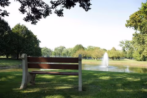 A wooden park bench on grass facing a pond with a fountain and trees in the background.