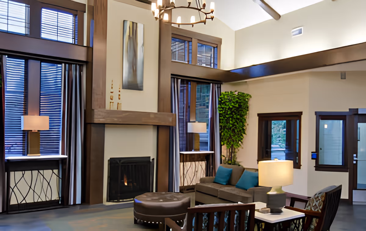 A cozy living room area with a high ceiling, large windows with curtains, a fireplace with a wooden mantle, a chandelier, a gray sofa with blue cushions, a round ottoman, a wooden armchair, two table lamps on side tables, and a tall green plant in the corner.