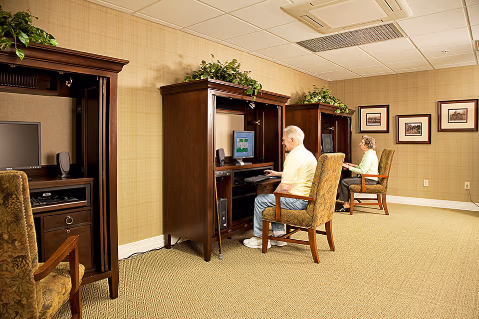 Two elderly individuals seated at separate wooden computer desks in a room with beige walls and carpet. Each desk has a computer monitor and keyboard, and there are framed pictures on the wall behind them. The room has a calm and organized atmosphere with plants placed on top of the desks.