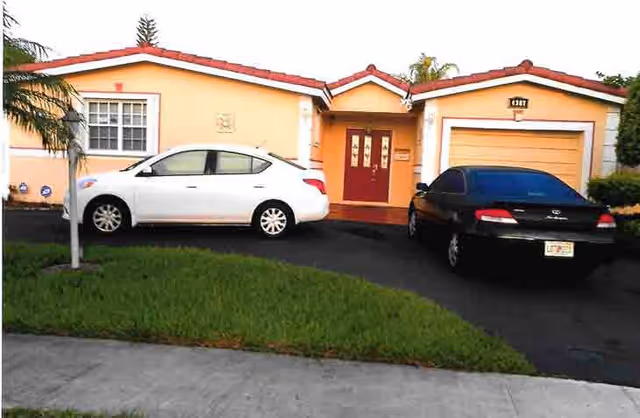 Front exterior view of a single-story house with a peach-colored facade, red roof tiles, a red front door, and a garage door. Two cars, one white and one black, are parked in the driveway. There is a small patch of grass and a palm tree near the sidewalk.