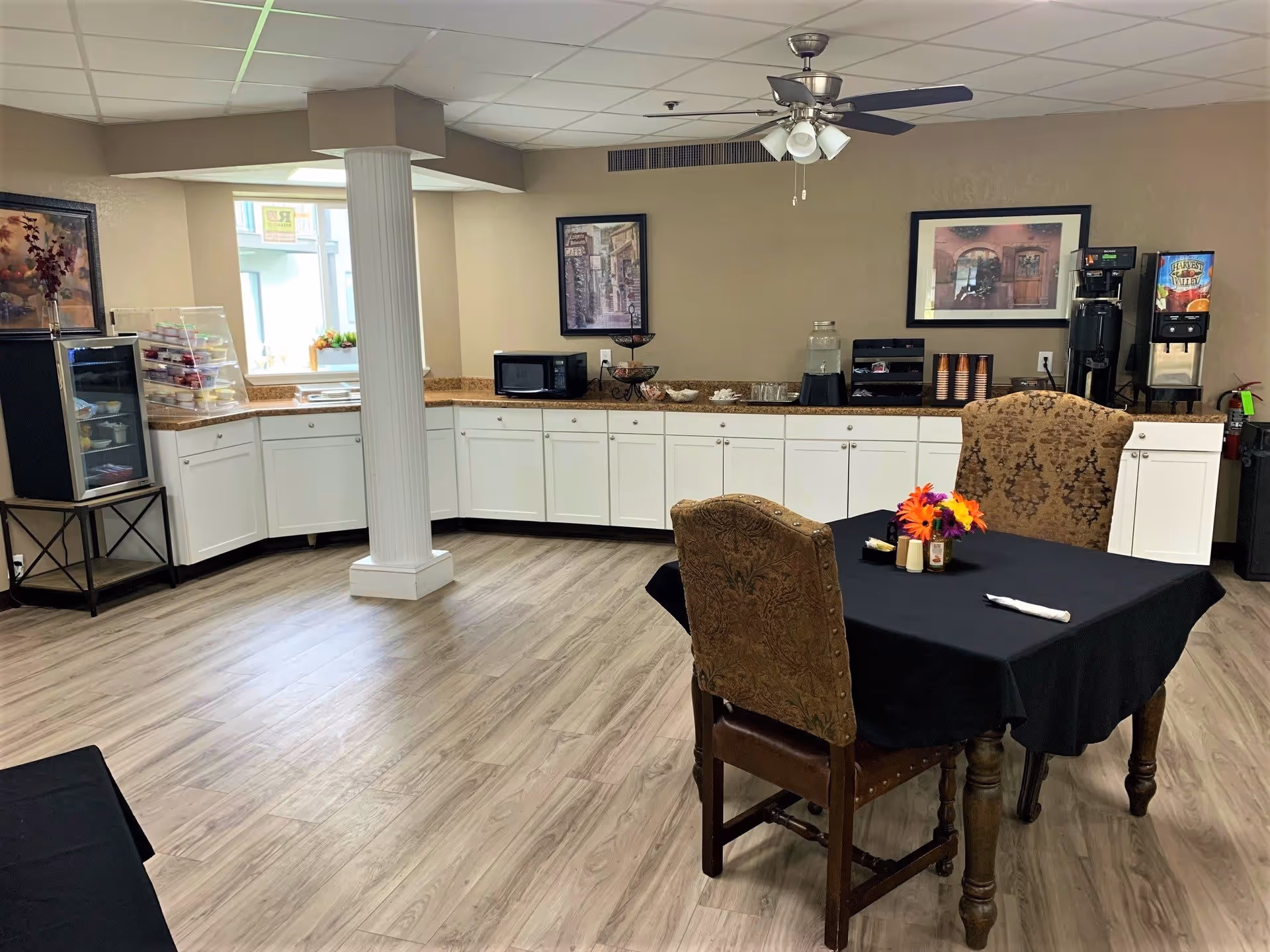 A cozy dining area in Southern Hills Retirement Community featuring a small table with two upholstered chairs covered in a patterned fabric. The table has a black tablecloth and a small floral centerpiece. In the background, there is a counter with white cabinets, a microwave, a coffee machine, a water dispenser, and a small refrigerator with snacks. Two framed pictures hang on the beige walls, and a ceiling fan with lights is mounted above.
