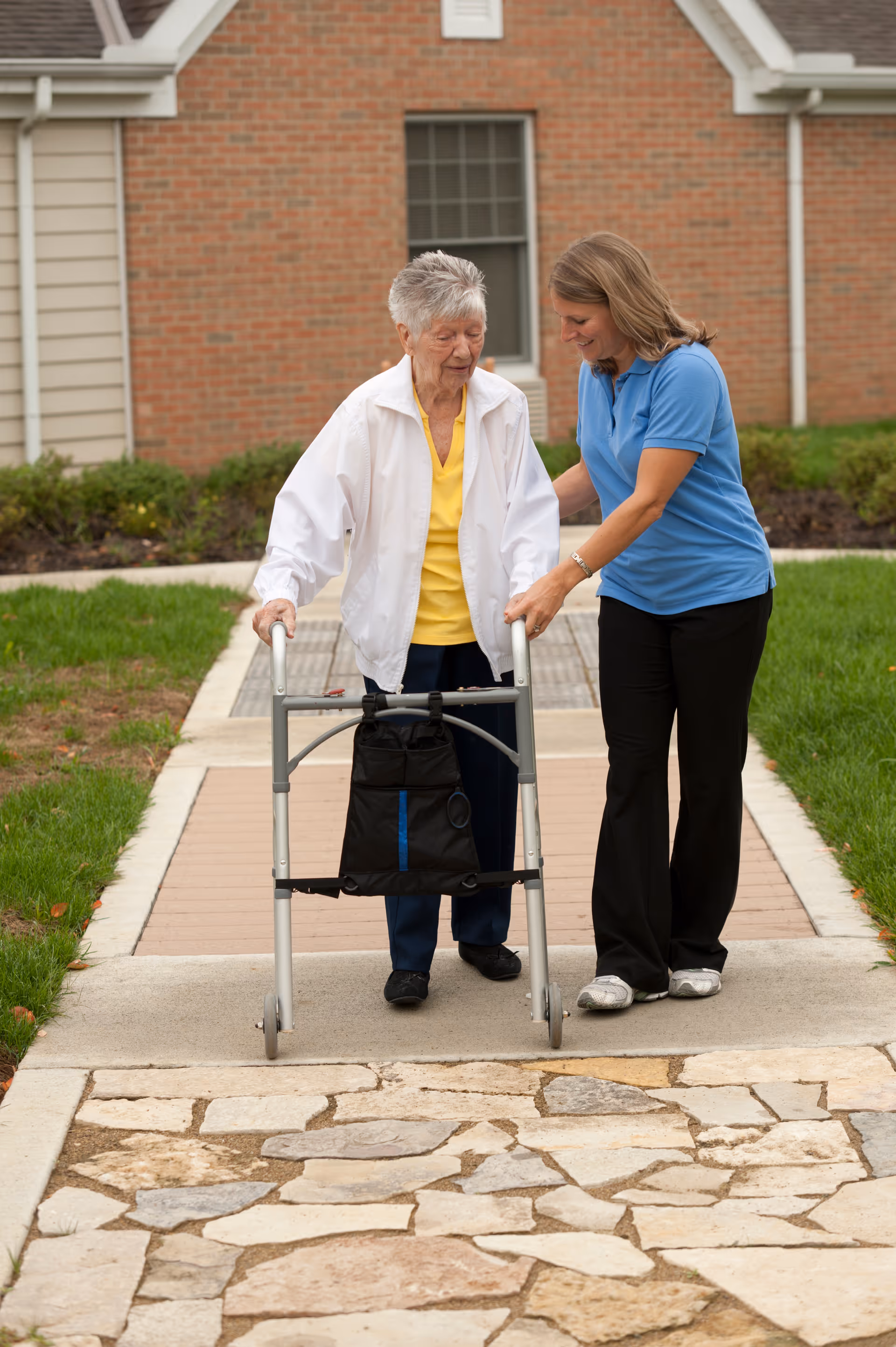 An elderly woman using a walker is being assisted by a caregiver wearing a blue shirt as they walk on a paved path outside a brick building with grass and shrubs nearby.
