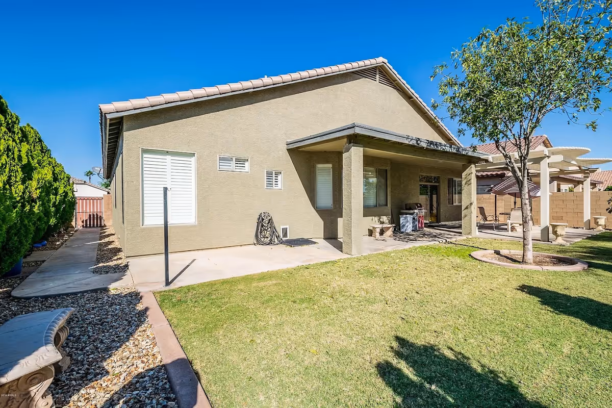 Backyard of a single-story stucco house showing a covered patio area, grassy lawn with a tree, and a paved side walkway.