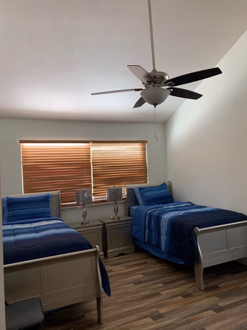 A bedroom with two single beds, each with blue bedding and pillows. Between the beds is a small nightstand with two decorative lamps. The room has wooden blinds covering the window and a ceiling fan with light fixture above. The floor is wood-patterned.