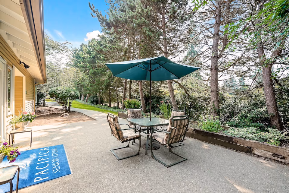 Outdoor patio area at Pacifica Dogwood Cottage with a glass table, four cushioned chairs, and a large green umbrella. The patio is adjacent to a yellow building with a blue mat that reads 'Pacifica Senior Living'. Surrounding the patio are trees and greenery under a partly cloudy sky.