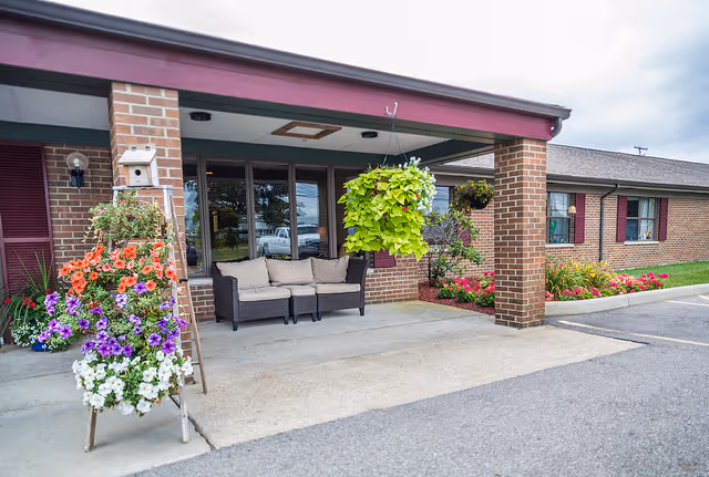 Entrance area of a brick building with a covered porch featuring a cushioned outdoor sofa. There are colorful flower arrangements on a stand and hanging plants near the entrance. The building has maroon window shutters and a parking area in front.