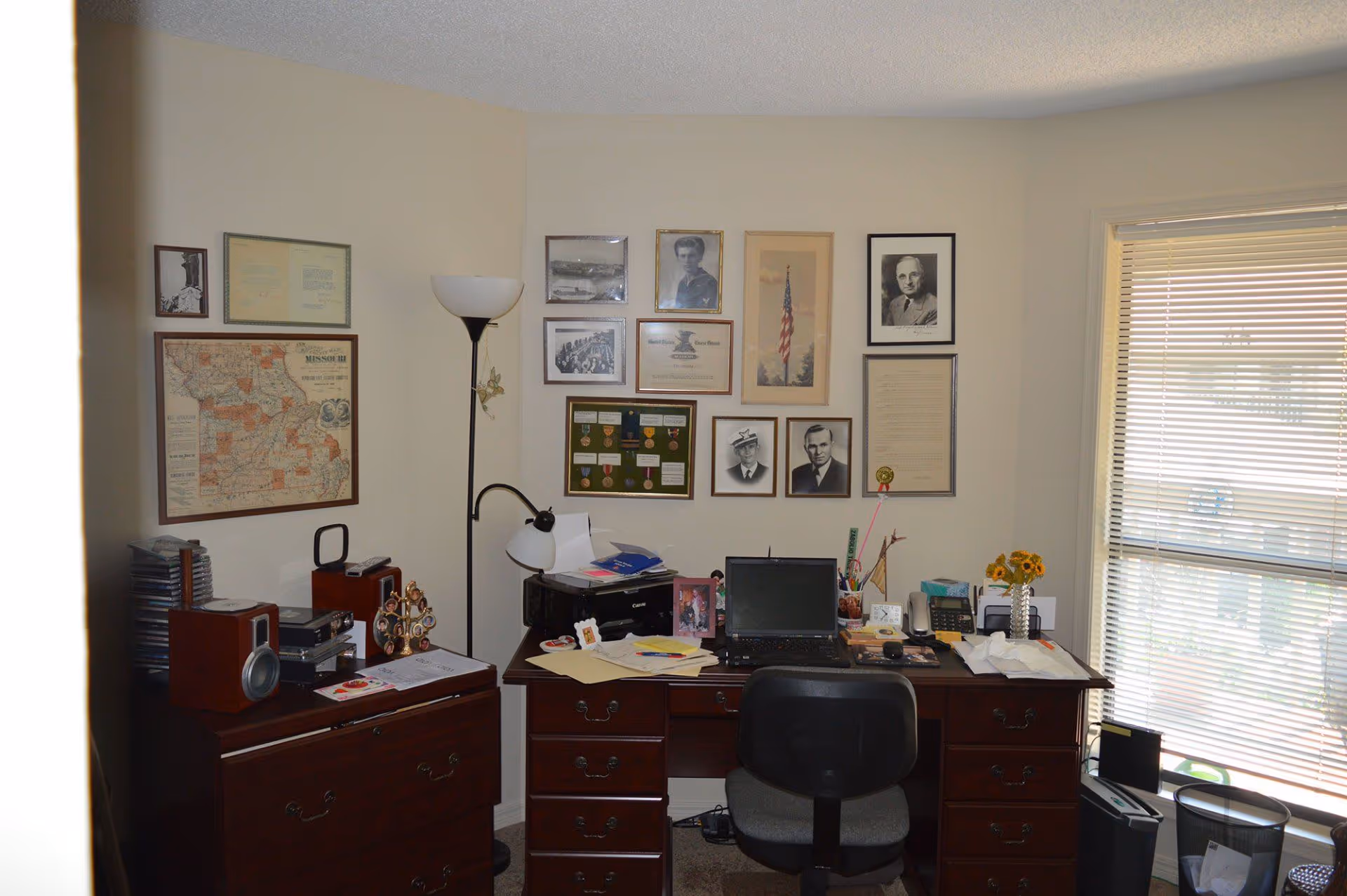 A home office setup with a wooden desk and chair. The desk holds a laptop, printer, papers, a telephone, and a vase with sunflowers. Behind the desk on the wall are framed photographs, certificates, and medals. To the left is a wooden filing cabinet with speakers and a CD rack on top. A tall floor lamp stands between the desk and filing cabinet. A window with blinds is on the right side of the room.