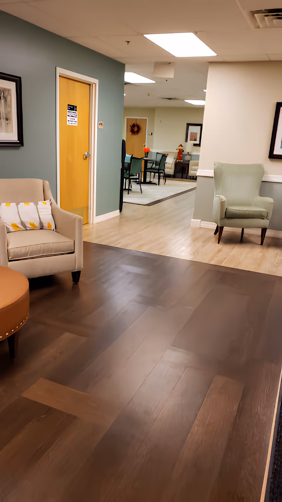 Interior view of a senior living facility lounge area with beige and green armchairs, a round ottoman, framed artwork on the walls, and a hallway leading to a dining area with tables and chairs. The floor transitions from dark wood to light wood, and the ceiling has recessed lighting.