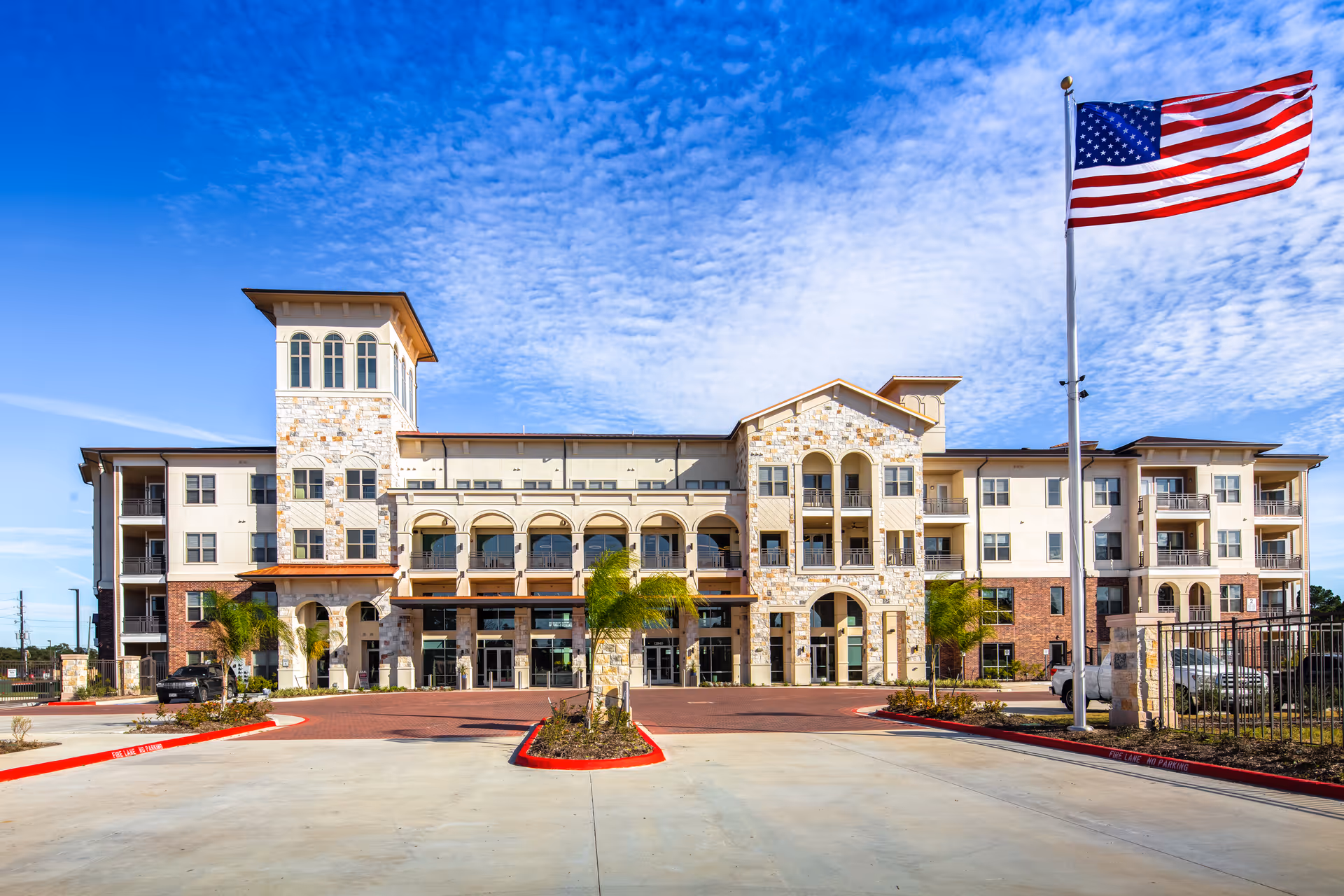 Front exterior view of Arella on Jones Active Senior Community building with a clear blue sky and an American flag on a flagpole in the foreground. The building features multiple floors, balconies, stone and brick facade, and arched architectural details.