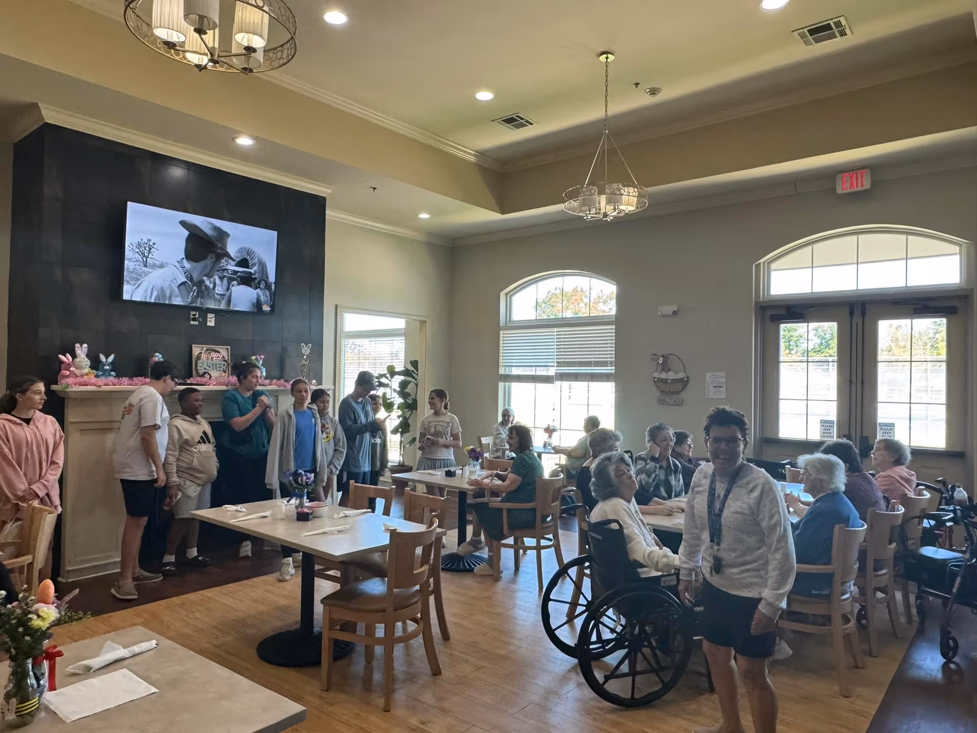 A group of elderly people sitting at tables in a well-lit common room with large windows. Several younger people stand near a fireplace decorated with Easter-themed items, and a TV above the fireplace shows a black and white movie. The room has wooden floors, modern light fixtures, and an exit door with windows.
