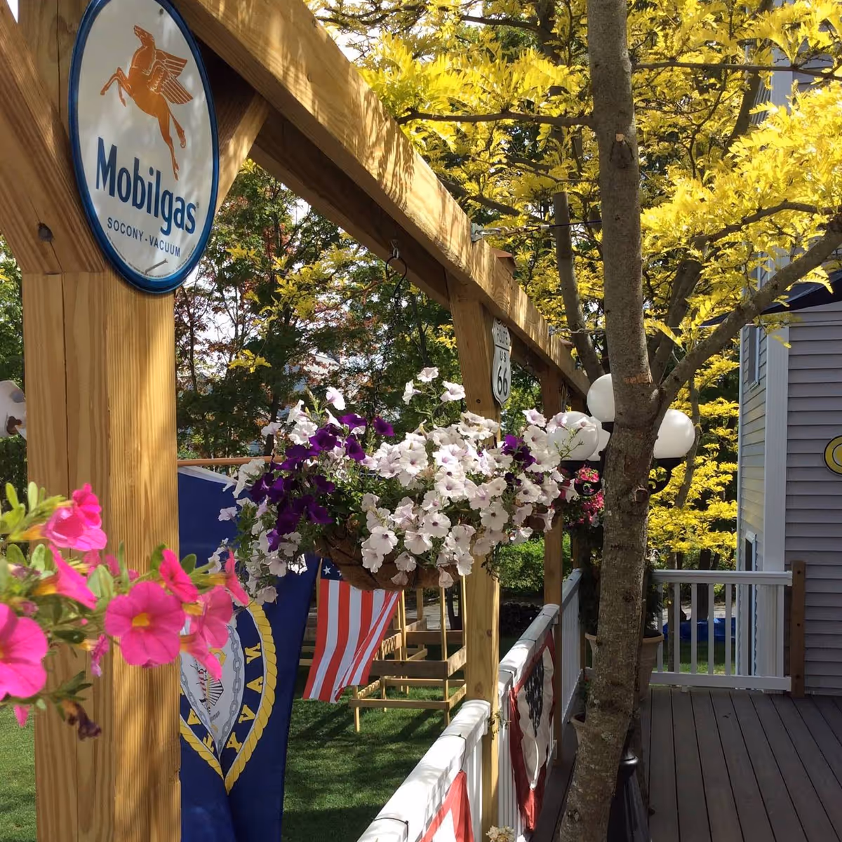 A wooden porch railing decorated with hanging flower baskets containing white and purple flowers, pink flowers in the foreground, American and Navy flags hanging, a tree growing through the porch, and a Mobilgas sign attached to the wooden beam.
