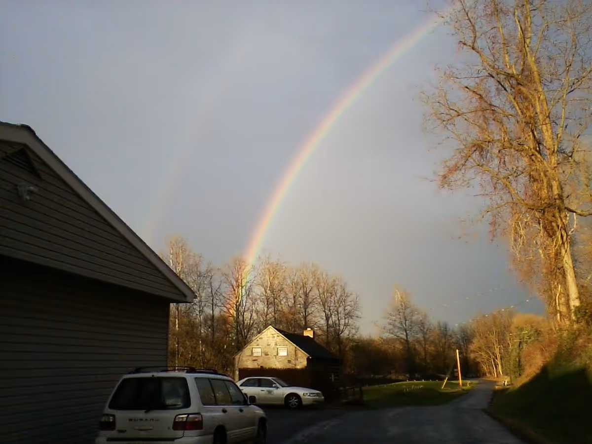 A double rainbow arcs over a rural scene with a small house, several parked cars, leafless trees, and a road leading into the distance under a cloudy sky.