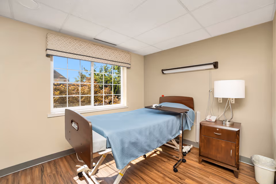 A small patient room with a single hospital bed covered in a blue blanket, a bedside table with a lamp and phone, and a window showing trees outside.