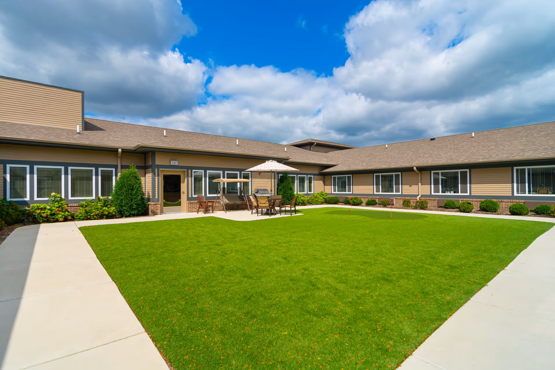 Outdoor courtyard area of a senior living facility named Harvester Place, featuring a well-maintained green lawn surrounded by a concrete walkway, patio furniture with an umbrella, and a building with multiple windows under a partly cloudy blue sky.