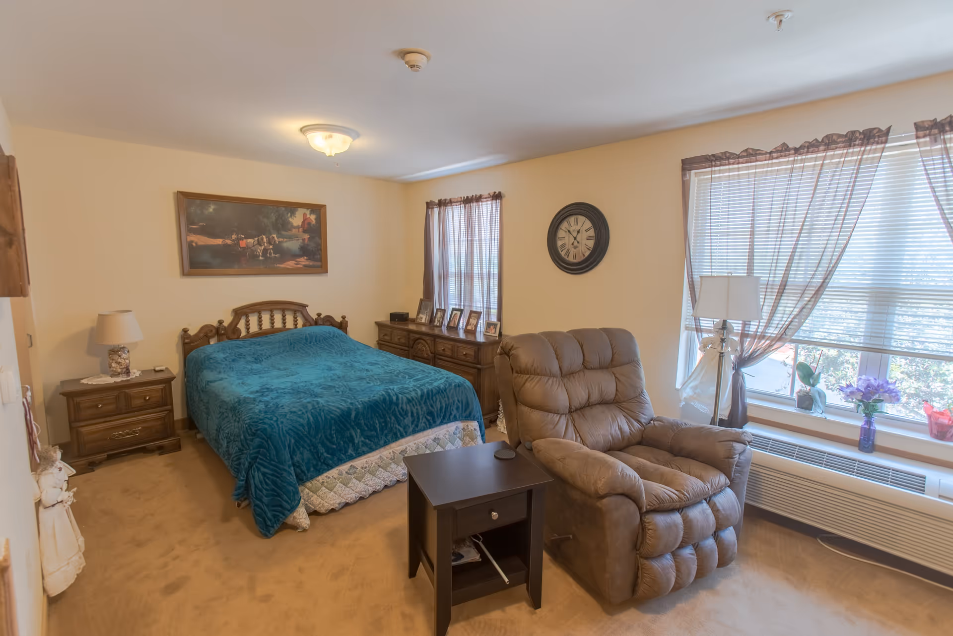 A furnished bedroom with a teal-covered bed, a brown recliner, wooden nightstands and dresser, and a large window with sheer curtains.