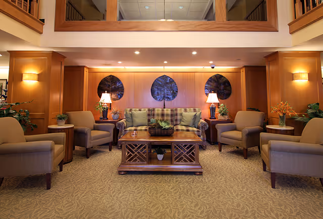A warm, well-lit lounge with a plaid sofa, armchairs, and a wooden coffee table arranged beneath recessed lighting and decorative wall art.