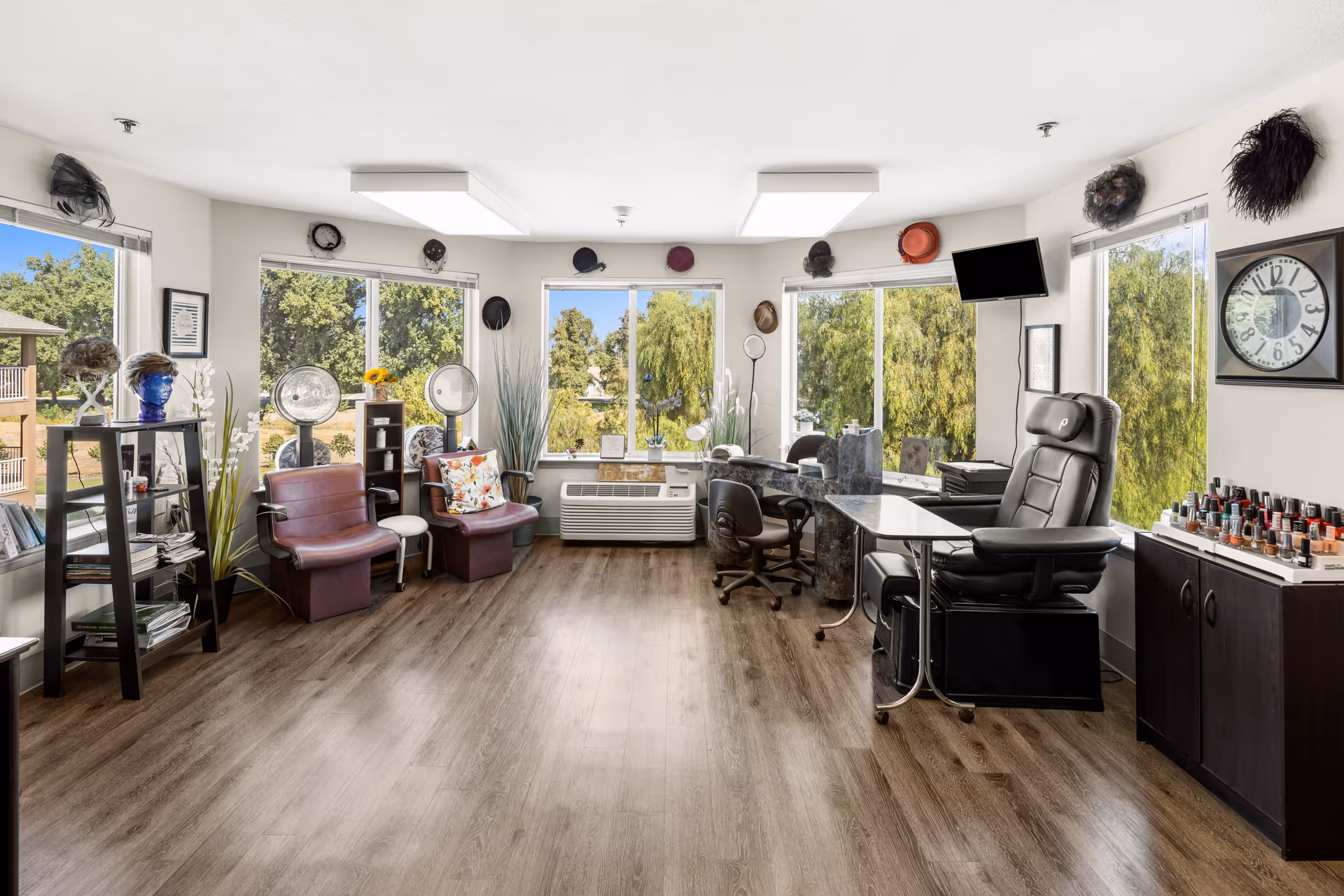 A bright and spacious salon room in a senior living community with large windows showing green trees outside. The room features two vintage hair dryers with maroon chairs, a manicure station with a black leather chair, a nail polish display, a wall clock, and various decorative hats mounted above the windows. The floor is wooden, and the walls are white, creating a clean and welcoming atmosphere.