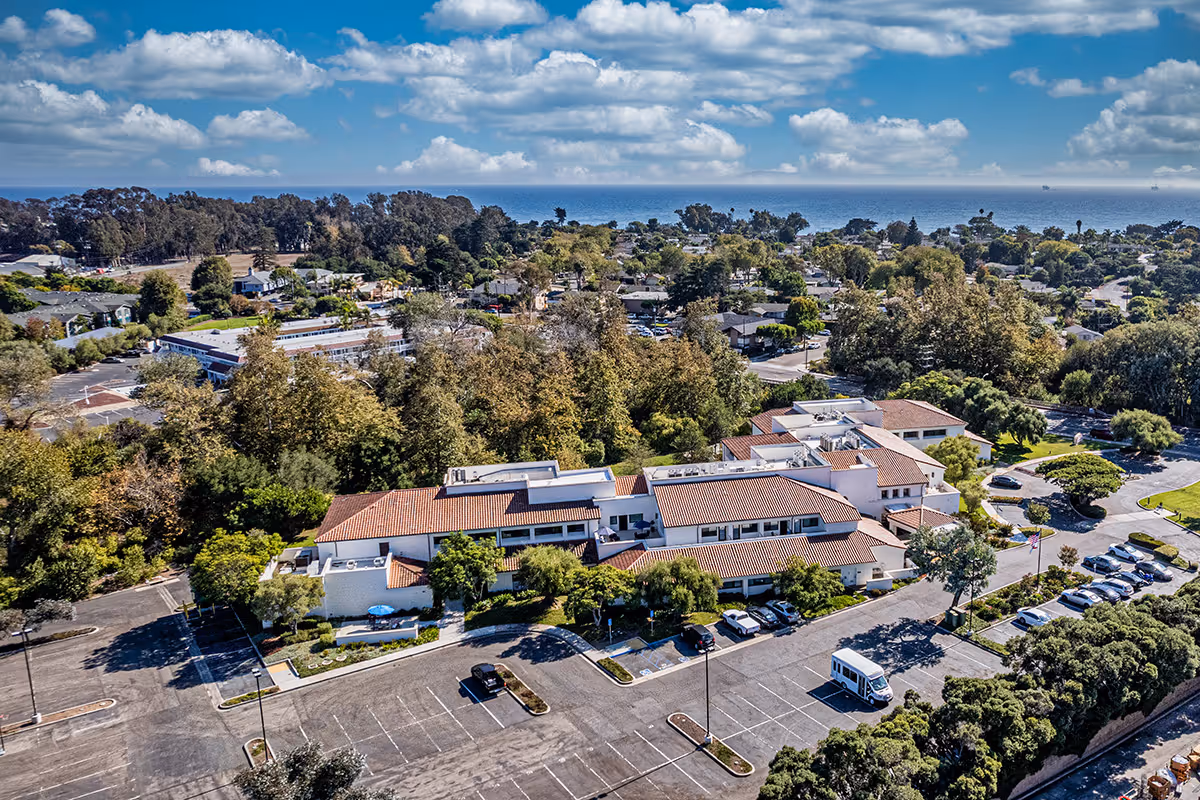 Aerial view of a terracotta-roofed senior living complex surrounded by trees, parking areas, and coastal scenery with the ocean visible on the horizon.